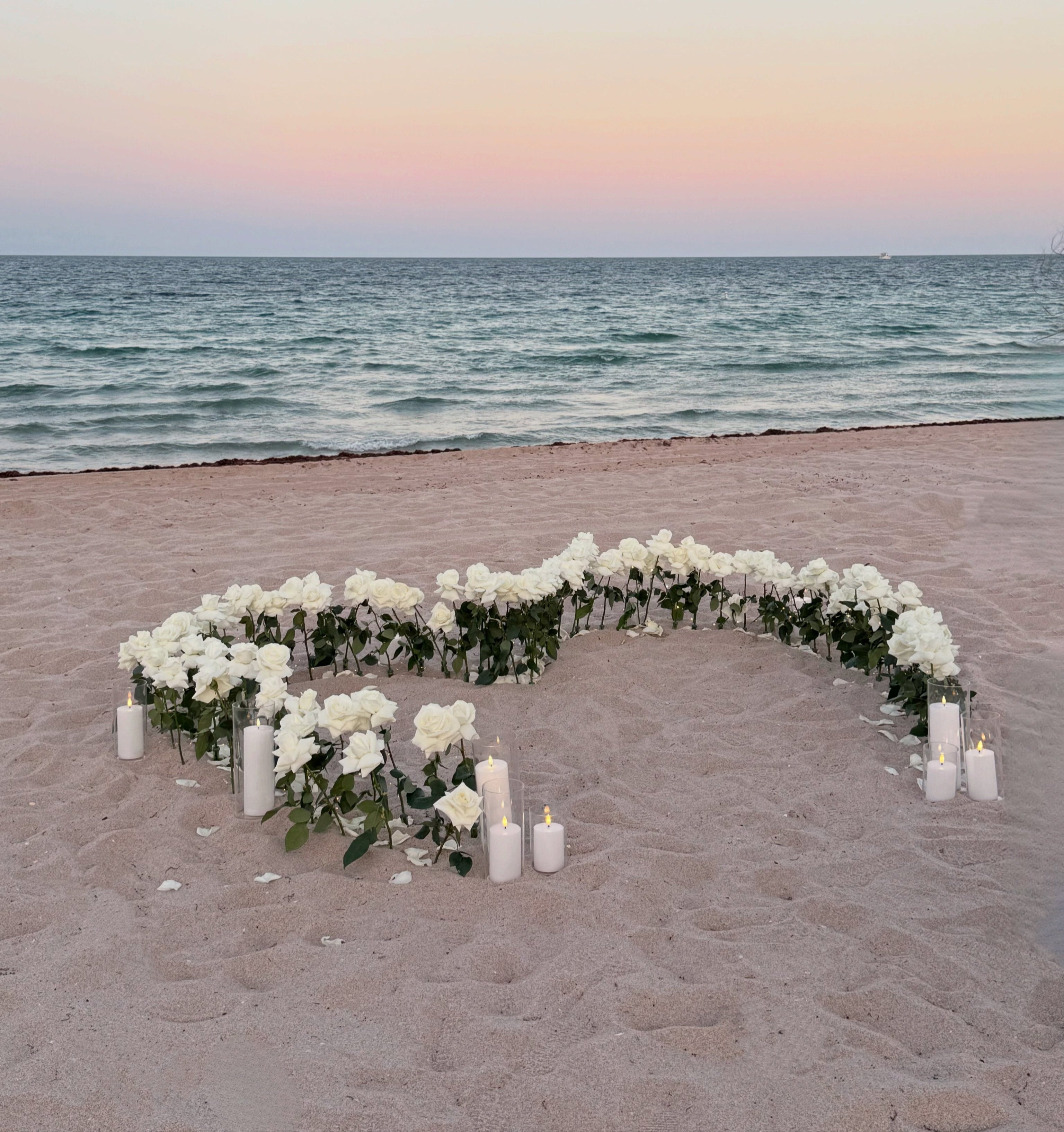 Heart-shaped arrangement of white flowers and candles on a sandy beach with ocean and sunset in the background
