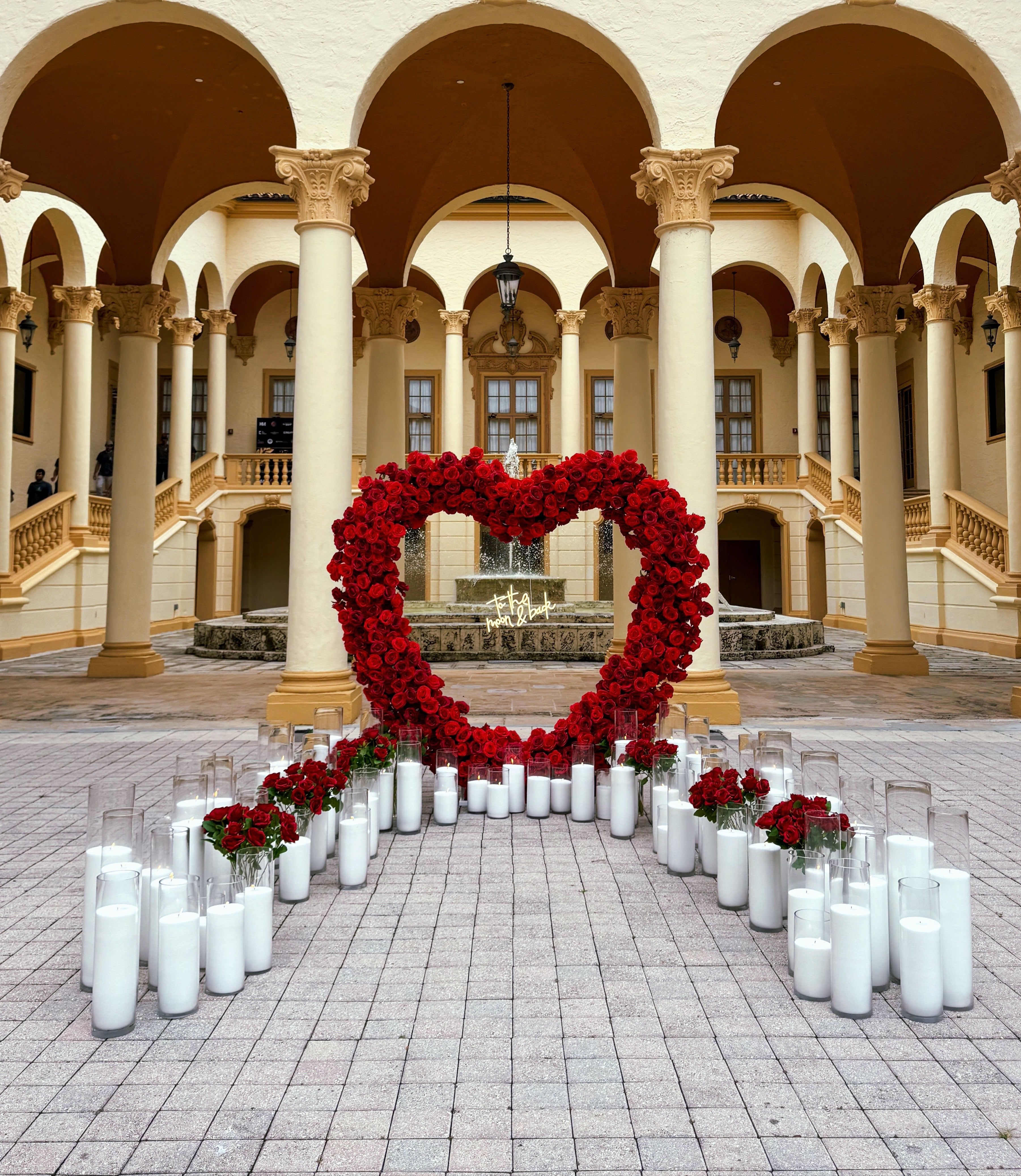 Heart-shaped arrangement of red flowers and white candles in a courtyard with arches and columns.