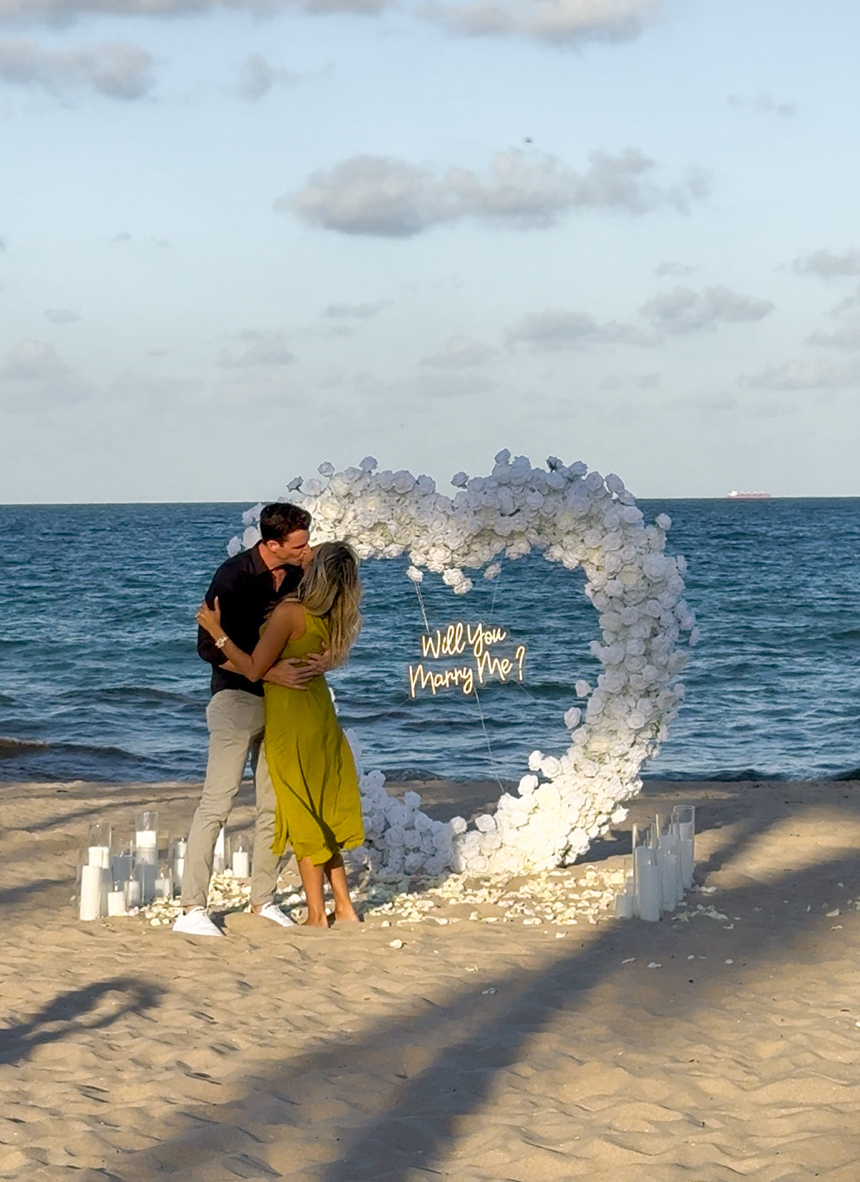 Couple embracing in front of a heart-shaped arch on a beach with 'Will You Marry Me?' proposal.