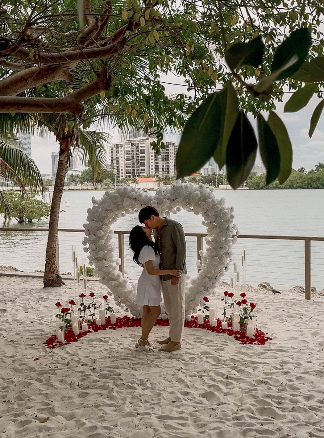 Couple kissing under a heart-shaped floral arch on a beach with palm trees and buildings in the background.