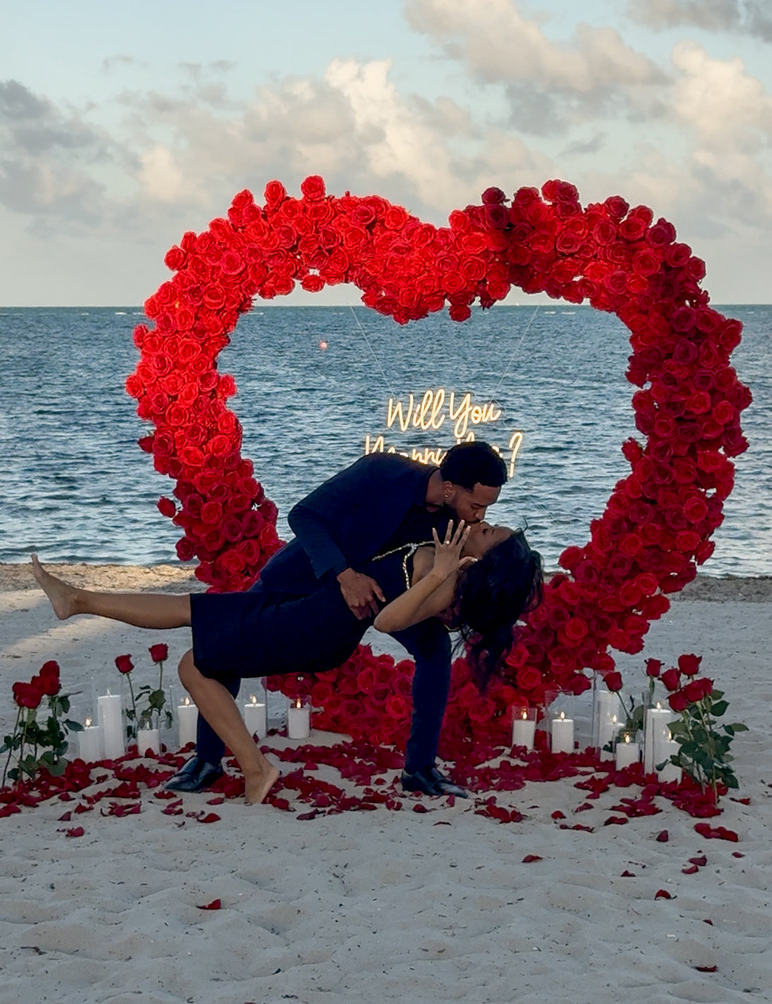 Couple in front of a heart-shaped arrangement of red flowers on a beach with 'Will you' proposal sign.