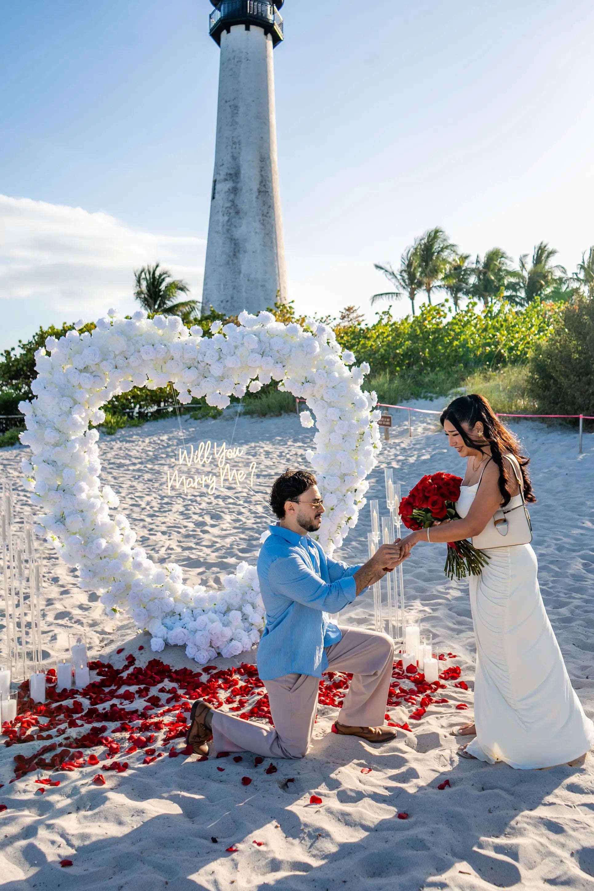 Man proposing to a woman in front of a heart-shaped floral arch on a beach with a lighthouse in the background.